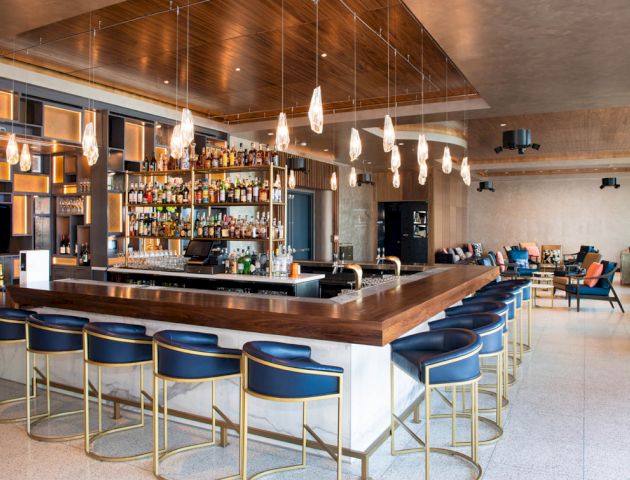 A modern bar features a wooden counter, blue bar stools, hanging lights, and a fully stocked bar. There is seating in an open, stylish space.