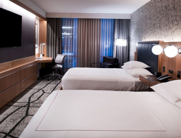 This image shows a modern hotel room with two double beds, a desk, a TV, and a window with curtains, featuring a stylish, contemporary design.