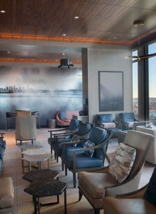 A modern lounge area with comfortable seating, large windows showcasing a waterfront view, and a wall mural of a cityscape in the background.