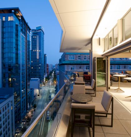 A modern balcony overlooking a cityscape at twilight, featuring outdoor seating and tables, adjacent to tall buildings and lit windows.