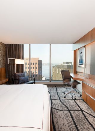 A modern hotel room with a large bed, desk, chair, TV, and a window with a city view. Minimalist decor with wood accents and artistic wallpaper.