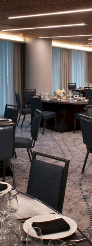 The image shows a banquet hall set for an event with round tables, chairs, and elegant place settings, including glassware and folded napkins. Curtains cover the windows.