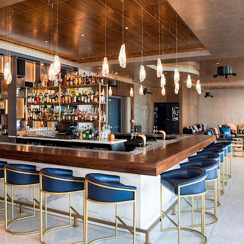 This image shows a modern bar with blue stools, pendant lights, and a well-stocked liquor shelf in a spacious, stylish indoor setting.