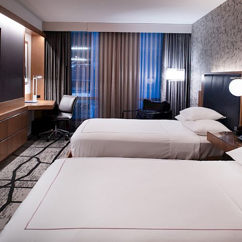 The image shows a modern hotel room with two neatly made beds, a flat-screen TV, a desk with a chair, and a large window with curtains.