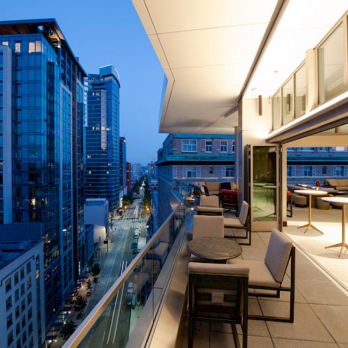 A rooftop terrace overlooking a city street lined with high-rise buildings, with modern outdoor seating and tables next to large glass windows.