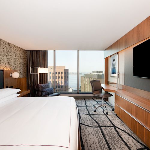 A modern hotel room features a large bed, wall-mounted TV, desk with chair, an armchair, and a window with a city view.