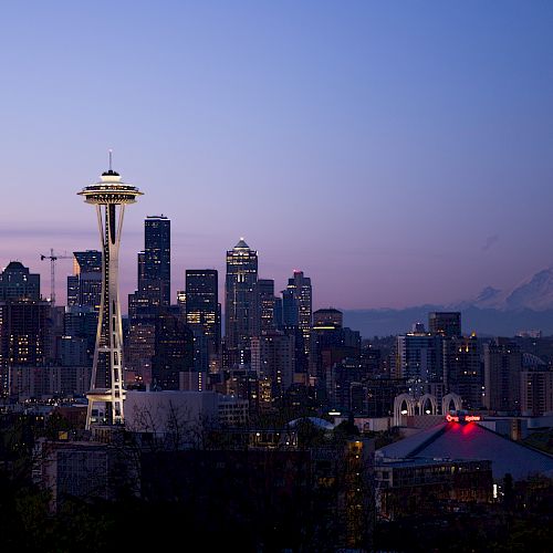 A nighttime view of Seattle with the illuminated Space Needle, surrounding cityscape, and Mount Rainier in the background under a twilight sky.
