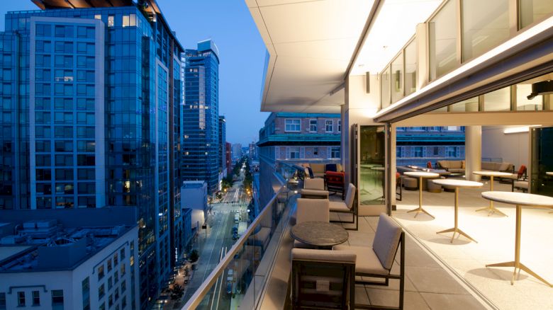 Modern high-rise buildings and a well-lit terrace with tables and chairs overlooking a city street at twilight.