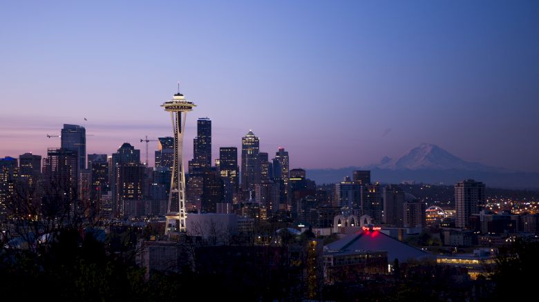 A cityscape at dusk featuring the Space Needle, various skyscrapers, and a distant mountain under a clear, fading sky.