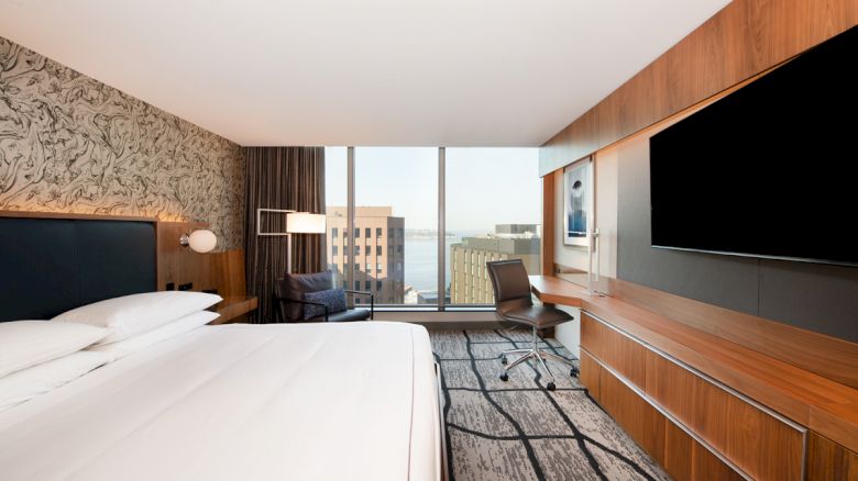 A modern hotel room with a large bed, a wall-mounted TV, a desk with a chair, and a view of city buildings through a large window ending the sentence.