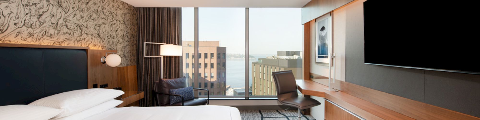 A modern hotel room with a large bed, desk, chair, flat-screen TV, and a window with a cityscape view, featuring contemporary design elements.